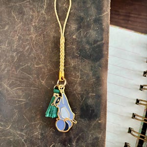 May include: A gold-colored charm with a blue and black cartoon character design, a green tassel, and a braided cord. The charm is hanging on a brown leather surface.
