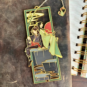 Puede incluir: Marcapáginas de metal dorado con una pareja sentada en una plataforma con fondo verde. El marcapáginas tiene una cadena con un dije en forma de corazón.