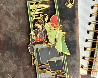 Pode incluir: Marca-páginas de metal dourado com um casal sentado em uma plataforma com fundo verde. O marca-páginas tem uma corrente com um amuleto em forma de coração.