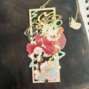 Könnte beinhalten: Goldener Metall-Lesezeichen mit einer farbenfrohen Illustration einer Frau in einem rot-weißen Kleid mit Blumenmuster. Das Lesezeichen hat eine Kette.