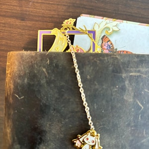 K&ouml;nnte beinhalten: Goldenes Lesezeichen mit einer Kette und einem wei&szlig;en Katzenanh&auml;nger. Das Lesezeichen ist in Form eines Rahmens mit einer Schriftrolle darin. Der Katzenanh&auml;nger hat rosa Ohren und einen rosa Schwanz.