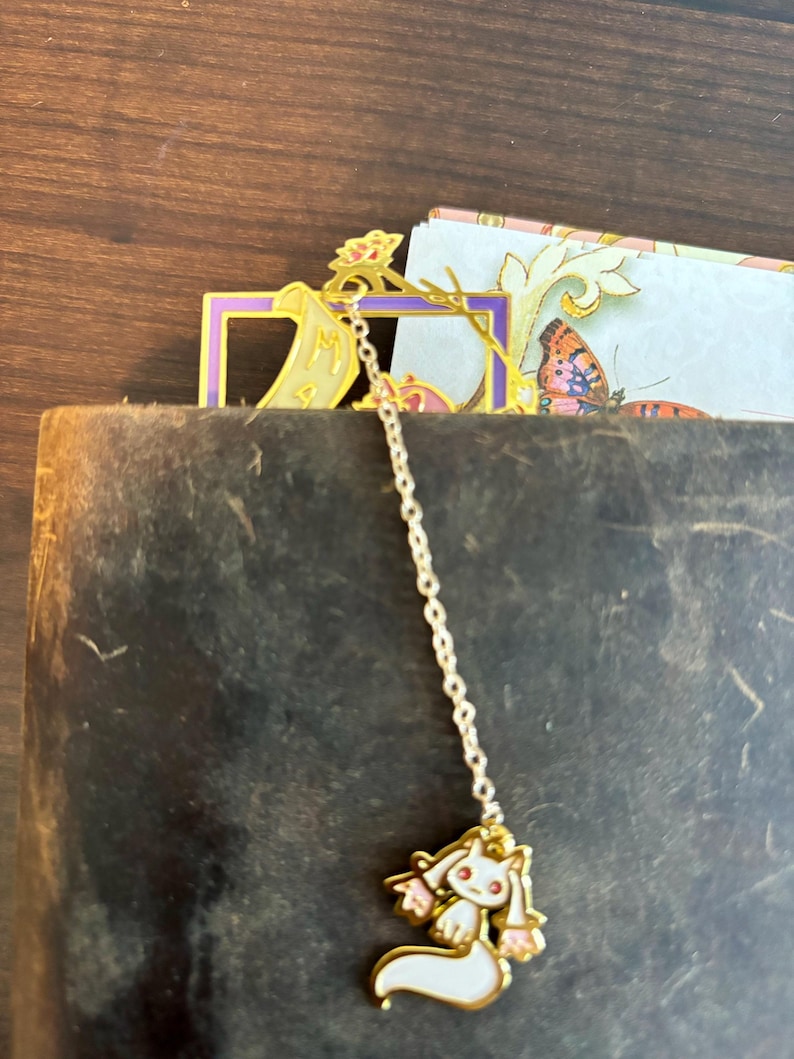 K&ouml;nnte beinhalten: Goldenes Lesezeichen mit einer Kette und einem wei&szlig;en Katzenanh&auml;nger. Das Lesezeichen ist in Form eines Rahmens mit einer Schriftrolle darin. Der Katzenanh&auml;nger hat rosa Ohren und einen rosa Schwanz.
