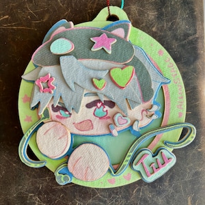 May include: A layered, decorative ornament featuring a cartoon character with blue hair and cat ears. The design includes pink, green, and blue accents, with star and heart details. The ornament has a string for hanging and the word "Till" is visible.