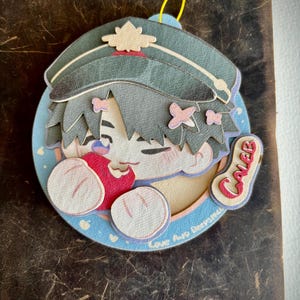 May include: A layered paper ornament depicting a cartoon character in a military hat. The character has blue hair, pink bows, and holds a red heart. The name "Caleb" is on a tag. The ornament has a blue background with the text "Love And Deepspace".