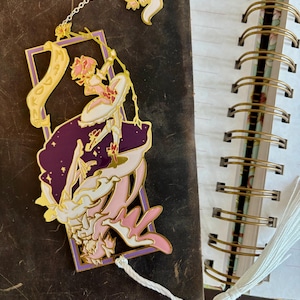K&ouml;nnte beinhalten: Ein goldfarbener Metall-Lesezeichen mit einer pink- und lilafarbenen Anime-Figur und einem wei&szlig;en Quasten. Das Lesezeichen hat eine Kette mit einem kleinen wei&szlig;en Katzenanh&auml;nger.