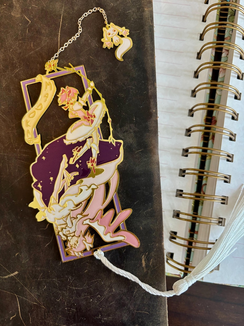 K&ouml;nnte beinhalten: Ein goldfarbener Metall-Lesezeichen mit einer pink- und lilafarbenen Anime-Figur und einem wei&szlig;en Quasten. Das Lesezeichen hat eine Kette mit einem kleinen wei&szlig;en Katzenanh&auml;nger.