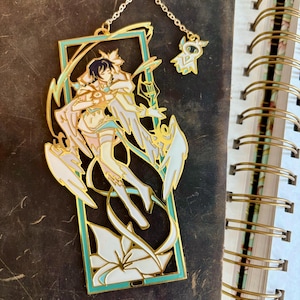 Puede incluir: Un marcapáginas de metal dorado con una ilustración detallada de una figura alada con una flor blanca. El marcapáginas está unido a una cadena dorada con un pequeño amuleto.