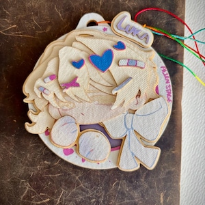 Op de afbeelding: Een gelaagde houten bedel met een stripfiguur met blond haar, blauwe harten en de naam "Luka". De bedel heeft een witte achtergrond met de tekst "Fallen Stage" en is geregen met kleurrijke koorden. De bedel ligt op een bruin lederen oppervlak.