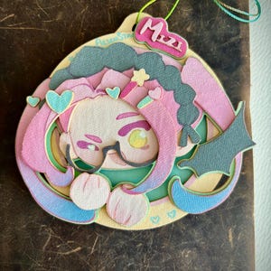 Op de afbeelding: Een gelaagd papieren kunstwerk met een stripfiguur met roze haar, een bril en een naamlabel met de tekst "Mizzi". Het kunstwerk bevat harten, sterren en een pastelkleurenpalet van roze, blauw en groen. Bovenop is een veelkleurig touwtje bevestigd.