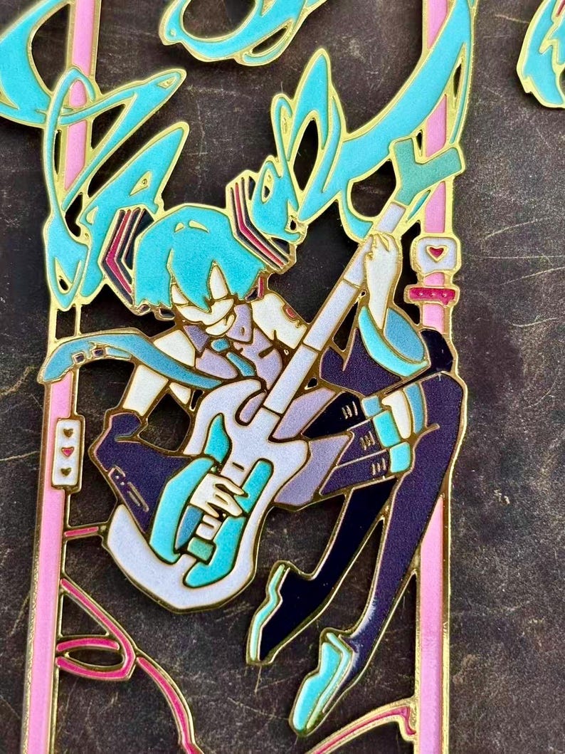 K&ouml;nnte beinhalten: Ein detaillierter Emaille-Pin mit einem Anime-Charakter mit t&uuml;rkisfarbenen Haaren, der eine wei&szlig;e und t&uuml;rkisfarbene Gitarre spielt. Der Pin hat goldfarbene Umrisse und rosa Akzente mit einem herzf&ouml;rmigen Detail. Der Charakter tr&auml;gt ein lila-t&uuml;rkisfarbenes Outfit.