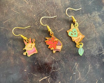 Pode incluir: Três pares de brincos pendurados dourados com desenhos de desenhos animados. Um par apresenta uma criatura rosa e roxa, outro uma flor vermelha e laranja, e o terceiro um personagem de rosto verde com um pingente de folha verde.