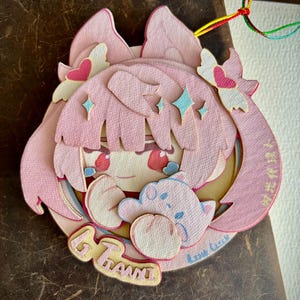 May include: A layered pink paper cutout depicting a cartoon character with pink hair, blue star accents, and a small cloud. The cutout has the text "67 Grams" and "Azure Circle". A multi-coloured string is attached.