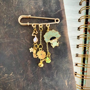 以下が含まれることがあります： 複数のチャームが付いたゴールドカラーの安全ピン。チャームには音符、花、小さな緑色のフィギュア、デイジー、ベルが含まれます。ピンは、濃い茶色のテクスチャ加工された表面に表示されています。