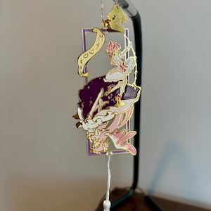 K&ouml;nnte beinhalten: Goldenes Lesezeichen mit einem lila und rosa emaillierten Design einer Frau mit einer Krone und einer Schriftrolle mit der Aufschrift "Kingdom". Das Lesezeichen hat einen wei&szlig;en Quasten.
