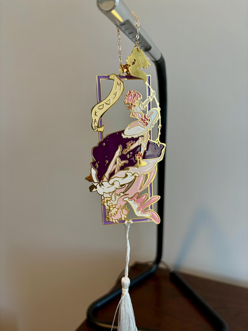 K&ouml;nnte beinhalten: Goldenes Lesezeichen mit einem lila und rosa emaillierten Design einer Frau mit einer Krone und einer Schriftrolle mit der Aufschrift "Kingdom". Das Lesezeichen hat einen wei&szlig;en Quasten.