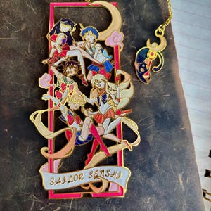 Könnte beinhalten: Goldfarbener Metall-Lesezeichen mit einem Design von Anime-Charakteren in farbenfrohen Outfits. Das Lesezeichen hat einen rosa rechteckigen Rahmen mit den Worten "SAILOR SENSHI" auf einem Banner. Eine goldene Kette verbindet einen Mond und einen Charakterkopf.