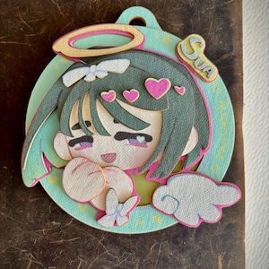 May include: A layered paper art piece depicting a cartoon character with green hair, pink hearts, and a halo. The character is set against a light blue background with the name "SUA" and a white cloud. It has a loop at the top.