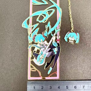 K&ouml;nnte beinhalten: Goldfarbener Metall-Lesezeichen mit einem stilisierten Anime-Charakter, der Gitarre spielt, mit t&uuml;rkisfarbenem Haar und Akzenten. Das Lesezeichen hat einen rosa Rand und einen kleinen Anh&auml;nger an einer Kette. Das Lesezeichen ist ca. 7,6 cm lang.