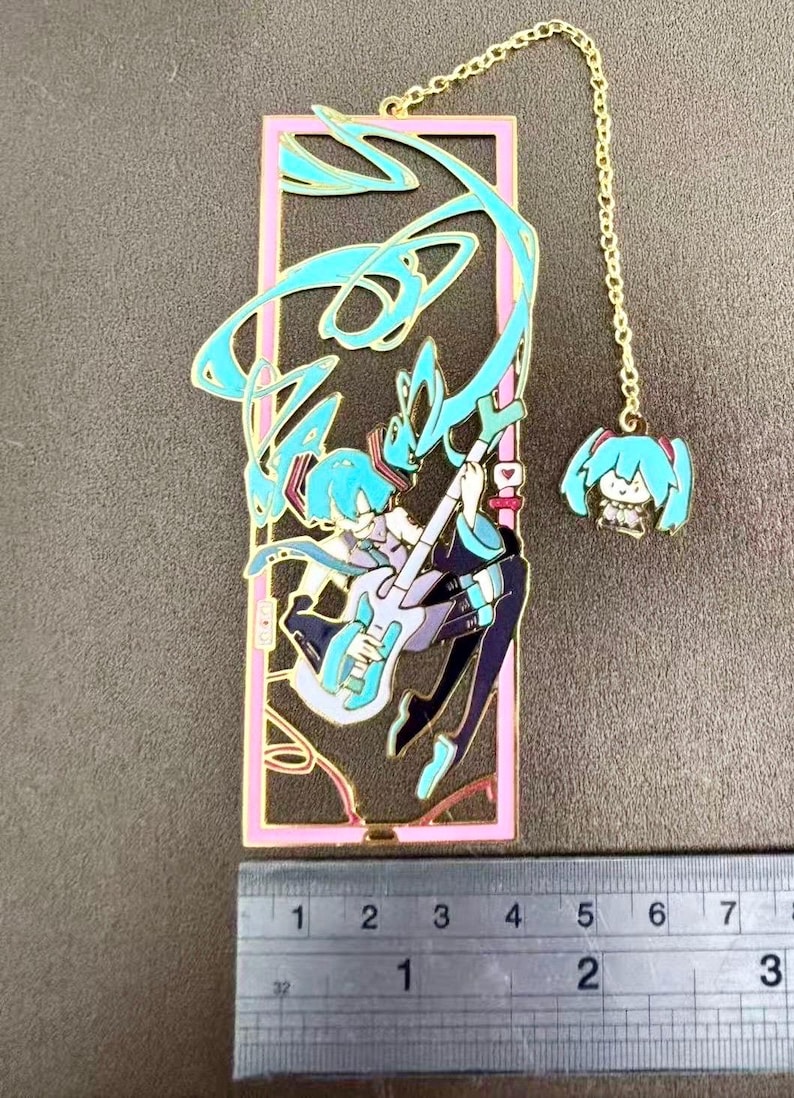 K&ouml;nnte beinhalten: Goldfarbener Metall-Lesezeichen mit einem stilisierten Anime-Charakter, der Gitarre spielt, mit t&uuml;rkisfarbenem Haar und Akzenten. Das Lesezeichen hat einen rosa Rand und einen kleinen Anh&auml;nger an einer Kette. Das Lesezeichen ist ca. 7,6 cm lang.