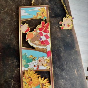 May include: A decorative bookmark with a gold chain and a charm. The bookmark features a whimsical design with a hamster, sunflowers, and a checkered pattern. The color palette includes brown, red, yellow, and blue.