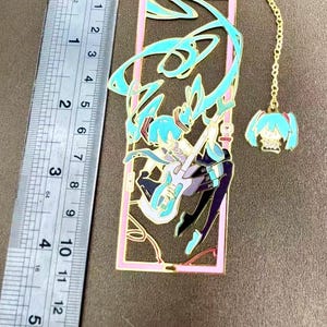 K&ouml;nnte beinhalten: Metall-Lesezeichen mit einem t&uuml;rkis-violetten Anime-Charakter, der Gitarre spielt, in einem rosa Rahmen. Das Lesezeichen hat eine goldene Kette mit einem Anh&auml;nger. Das Lesezeichen ist etwa 13 cm lang.