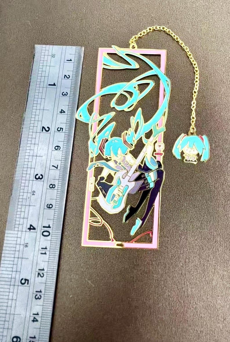 K&ouml;nnte beinhalten: Metall-Lesezeichen mit einem t&uuml;rkis-violetten Anime-Charakter, der Gitarre spielt, in einem rosa Rahmen. Das Lesezeichen hat eine goldene Kette mit einem Anh&auml;nger. Das Lesezeichen ist etwa 13 cm lang.