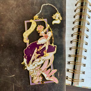 K&ouml;nnte beinhalten: Goldener Metall-Lesezeichen mit einer pinkfarbenen und wei&szlig;en Anime-Figur mit einem Zauberstab, die auf einer lila und wei&szlig;en Wolke steht. Die Figur h&auml;lt ein Banner mit der Aufschrift "Madoka".
