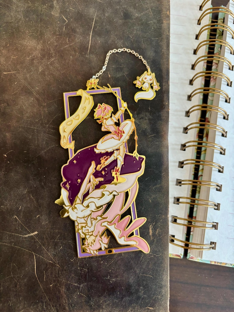 K&ouml;nnte beinhalten: Goldener Metall-Lesezeichen mit einer pinkfarbenen und wei&szlig;en Anime-Figur mit einem Zauberstab, die auf einer lila und wei&szlig;en Wolke steht. Die Figur h&auml;lt ein Banner mit der Aufschrift "Madoka".