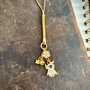 Puede incluir: Un dije de teléfono dorado con un lazo de cordón trenzado. El dije presenta una figura blanca y azul con forma de búho, una flor y una pequeña campana. El fondo es una superficie texturizada de color marrón oscuro.