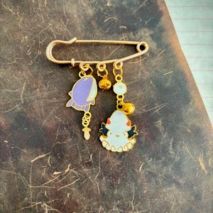 May include: A gold-coloured safety pin with two dangling charms. One charm is a purple and white whale, and the other is a white and black ghost-like figure. The charms are connected with gold-coloured chains and small gold beads.