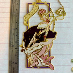 K&ouml;nnte beinhalten: Goldener Metall-Lesezeichen mit einer Comicfigur mit rosa Haaren und einem wei&szlig;en Kleid, die eine Schriftrolle mit dem Wort "DO" h&auml;lt. Die Figur ist von Sternen und einem violetten Rahmen umgeben. Ein kleiner wei&szlig;er Hasenanh&auml;nger h&auml;ngt am Lesezeichen.