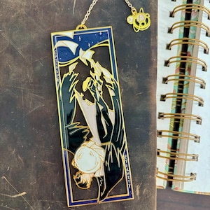 Może przedstawiać: Złoty metalowy zakładka do książki z niebiesko-czarną ilustracją postaci. Zakładka ma łańcuszek z zawieszką. Słowo "Eternity" jest napisane pionowo z boku.