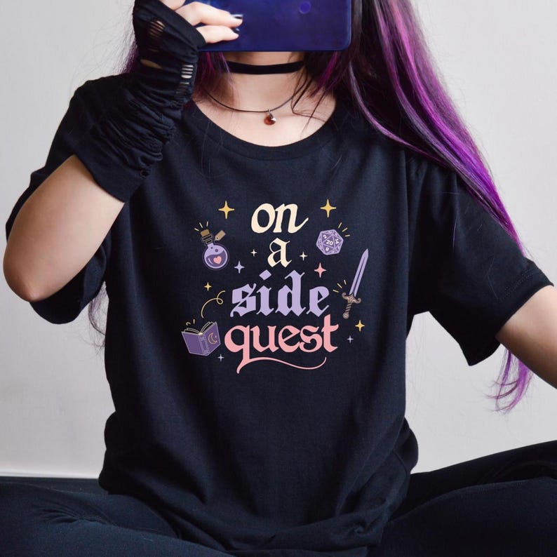 May include: Black t-shirt with the phrase "on a side quest" in a whimsical font, accompanied by fantasy-themed illustrations. The design includes a potion bottle, a die, a sword, and an open book. The shirt is short-sleeved.