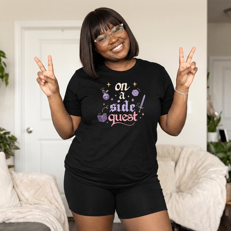 May include: Black t-shirt with the text "on a side quest" in a fantasy-themed design. The design includes a potion bottle, dice, a sword, and a book. The person is wearing black shorts and glasses.