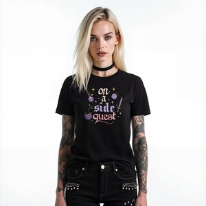 May include: Black t-shirt with the text "on a side quest" in a whimsical font, accompanied by fantasy-themed illustrations. The design includes dice, a potion bottle, and a sword. The model is wearing black jeans and a black choker.