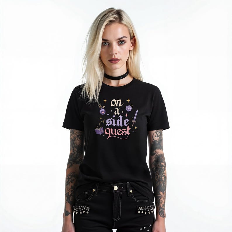 May include: Black t-shirt with the text "on a side quest" in a whimsical font, accompanied by fantasy-themed illustrations. The design includes dice, a potion bottle, and a sword. The model is wearing black jeans and a black choker.