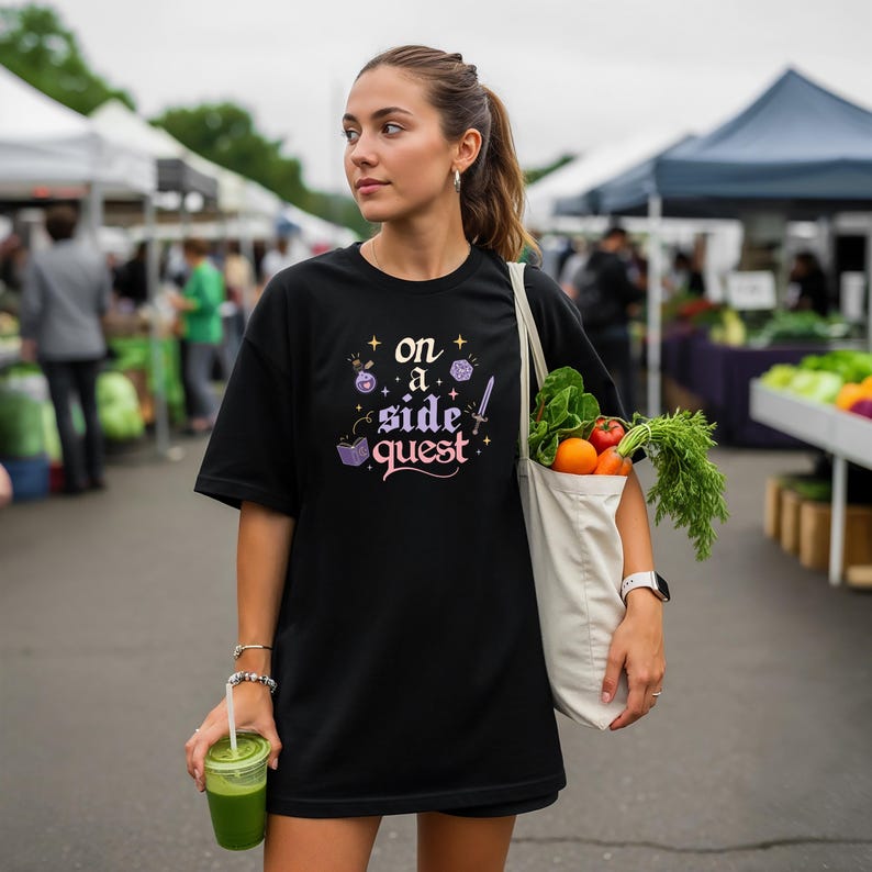 May include: A black t-shirt with the text "on a side quest" in a whimsical font, accompanied by fantasy-themed graphics. The person is carrying a tote bag filled with fresh produce and a green drink.