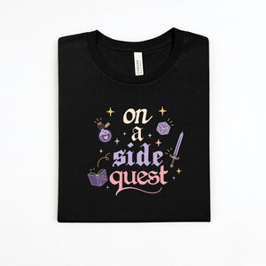 May include: Black t-shirt with the words "on a side quest" in a whimsical font, accompanied by fantasy-themed illustrations. The design includes a potion bottle, a die, a sword, and a book.