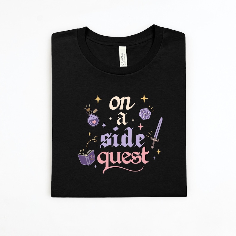 May include: Black t-shirt with the words "on a side quest" in a whimsical font, accompanied by fantasy-themed illustrations. The design includes a potion bottle, a die, a sword, and a book.