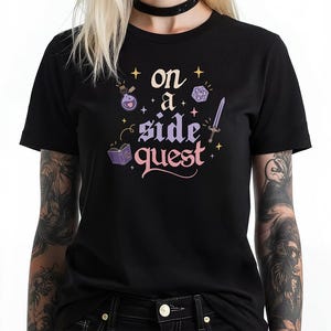 May include: Black t-shirt with the phrase "on a side quest" in a whimsical font. The design includes illustrations of a potion bottle, a book, a sword, and a die, all in a fantasy theme.