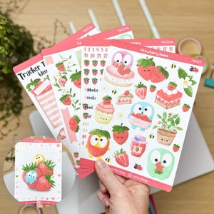 Könnte beinhalten: Eine Hand hält mehrere Stickerbögen mit Erdbeermotiv. Die Sticker zeigen Illustrationen von Erdbeeren, Kuchen und Eulen. Der Text enthält "Tracker Ideas" und "strawberry kisses". Die Sticker sind rosa, rot, grün und weiß.