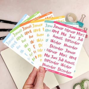 Könnte beinhalten: Nahaufnahme einer Hand, die einen Fächer aus bunten Aufklebern hält. Jedes Blatt hat einen andersfarbigen Rand und zeigt die Monatsnamen in einer handschriftlichen Schriftart. Aufkleber für die Planung.