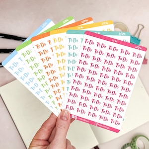 Op de afbeelding: Een hand houdt een waaier van kleurrijke plannerstickers vast. Elke sticker heeft de woorden "To Do" in een handschrift. De stickers zijn in tinten blauw, oranje, geel, groen en roze. De merknaam "natika design" is zichtbaar.