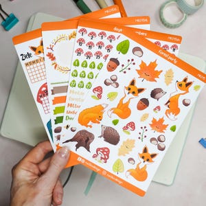 Könnte beinhalten: Eine Sammlung von Stickerbögen mit orangefarbenem Rand, die herbstliche Illustrationen zeigen. Designs umfassen Füchse, Pilze, Blätter, Eicheln und Kalenderelemente. Der Text enthält "forest party" und Kalendertage.