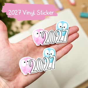 May include: Two vinyl stickers featuring the text 2027 in a bold black font. Each sticker displays a small pink creature and a small blue creature with large white eyes. The stickers have a white border and a watercolor texture on the characters.