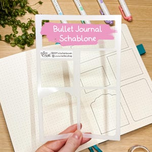 Könnte beinhalten: Eine halbtransparente Kunststoffschablone für Bullet Journals mit verschiedenen rechteckigen und dekorativen Ausschnitten. Die Schablone enthält den aufgedruckten Text SB001 Wochenboxen und www.natiko.shop. Sie liegt auf einer gepunkteten Notizbuchseite.