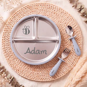 May include: A stainless steel divided plate with a light blue rim, a bee graphic, and the name "Adam" in green script. Includes a matching fork and spoon with light blue handles. The plate is approximately 8 inches in diameter.