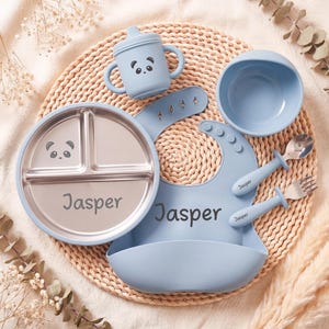 May include: A baby feeding set in light blue, featuring a divided plate with a panda face, a sippy cup, a bowl, a bib, and a spoon and fork. The name "Jasper" is printed on the plate and bib.