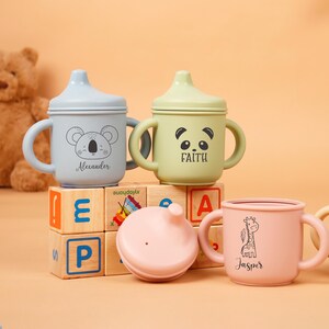 Puede incluir: Una colecci&oacute;n de vasos con boquilla para beb&eacute;s en colores pastel, cada uno con una ilustraci&oacute;n de animal &uacute;nica y el nombre de un ni&ntilde;o. Las tazas tienen asas y tapas, y est&aacute;n dispuestas sobre bloques de madera. Un oso de peluche y banderas decorativas est&aacute;n en el fondo.