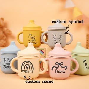 Puede incluir: Una colecci&oacute;n de coloridas tazas para beb&eacute;s con asas y tapas. Cada taza presenta un dise&ntilde;o diferente, incluyendo una cebra, un elefante, un panda, una corona, un arco&iacute;ris y un lazo. Las tazas son en tonos amarillo, gris, rosa, verde y azul.